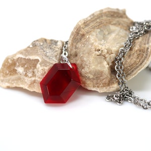 May include: A red gemstone pendant necklace with a silver chain. The pendant is a faceted, hexagonal shape and is resting on a piece of rock.