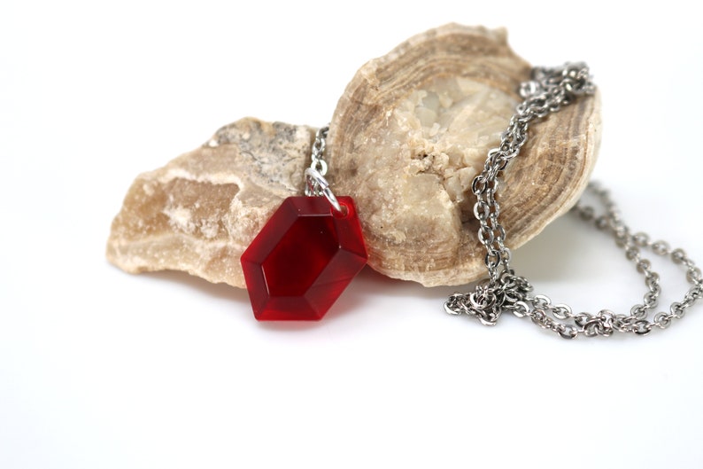 May include: A red gemstone pendant necklace with a silver chain. The pendant is a faceted, hexagonal shape and is resting on a piece of rock.