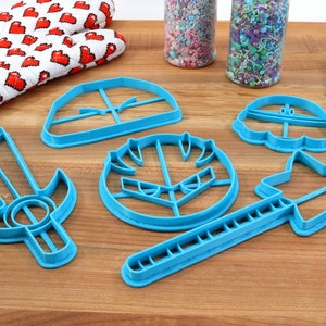 May include: A set of six blue plastic cookie cutters shaped like a sword, shield, star, mushroom, and two different hats. The cutters are on a wooden surface.
