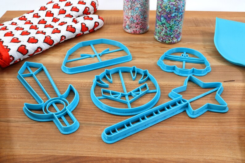 May include: A set of six blue plastic cookie cutters shaped like a sword, shield, star, mushroom, and two different hats. The cutters are on a wooden surface.