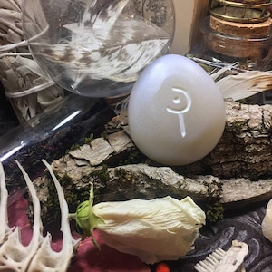May include: A white, smooth, egg-shaped stone with a white symbol carved into it. The stone is resting on a piece of bark with other natural objects around it.