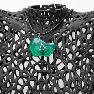 May include: A green pendant necklace with a silver chain. The pendant is shaped like a crescent moon with a stylized symbol inside. The necklace is displayed on a black mannequin.