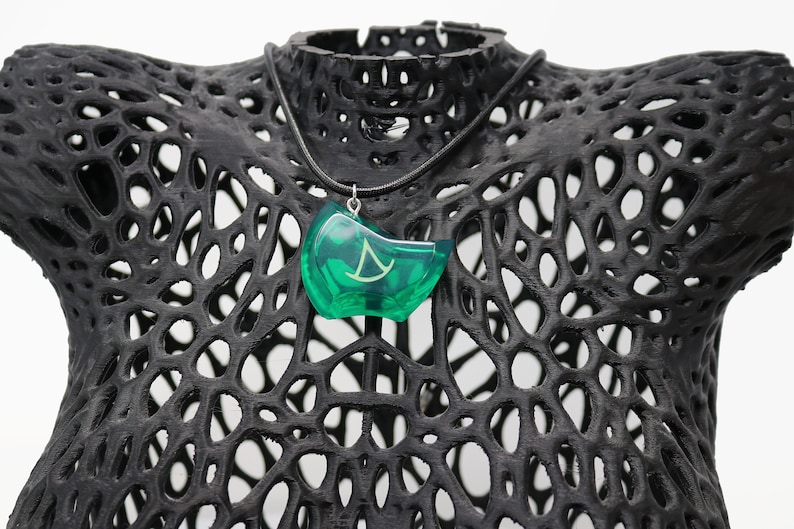 May include: A green pendant necklace with a silver chain. The pendant is shaped like a crescent moon with a stylized symbol inside. The necklace is displayed on a black mannequin.