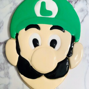 May include: A decorated sugar cookie in the shape of a cartoon character's face. The cookie features a green hat with a white circle and a green "L", black mustache, and large black eyes. The cookie is beige with a glossy finish.