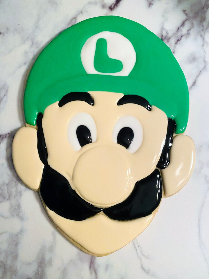 May include: A decorated sugar cookie in the shape of a cartoon character's face. The cookie features a green hat with a white circle and a green "L", black mustache, and large black eyes. The cookie is beige with a glossy finish.