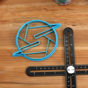 May include: A blue plastic cookie cutter shaped like a stylized letter S with a circle around it. The cutter is on a wooden surface with a black ruler.