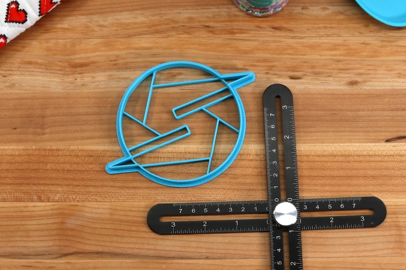 May include: A blue plastic cookie cutter shaped like a stylized letter S with a circle around it. The cutter is on a wooden surface with a black ruler.