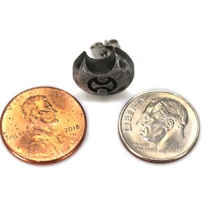 May include: A silver earring featuring a textured, crescent moon design with a small, black symbol at its centre. The earring is displayed alongside a US penny and a US dime to illustrate its size.
