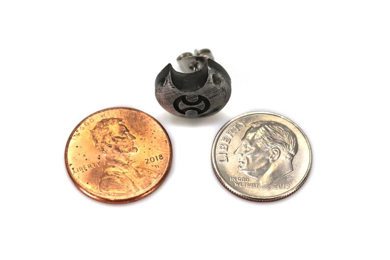 May include: A silver earring featuring a textured, crescent moon design with a small, black symbol at its centre. The earring is displayed alongside a US penny and a US dime to illustrate its size.