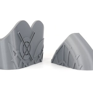 May include: Two gray, 3D-printed decorative pieces. One is a curved shape with a geometric pattern, and the other is a taller, shield-like shape with a circular design and cross. Both are on a white background.