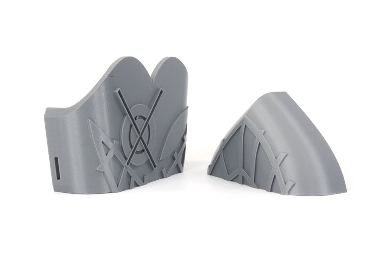 May include: Two gray, 3D-printed decorative pieces. One is a curved shape with a geometric pattern, and the other is a taller, shield-like shape with a circular design and cross. Both are on a white background.