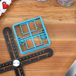 May include: A blue plastic cookie cutter shaped like a handheld video game console. The cutter has a square shape with a rectangular screen and buttons on the side. The cutter is on a wooden surface with a black ruler and a blue spatula.