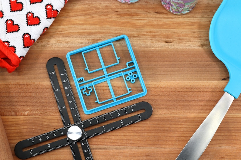 May include: A blue plastic cookie cutter shaped like a handheld video game console. The cutter has a square shape with a rectangular screen and buttons on the side. The cutter is on a wooden surface with a black ruler and a blue spatula.