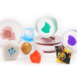 May include: A collection of colorful glass and resin plugs with various designs, including a pink, green, purple, and gold plug. The plugs are arranged on a white surface.