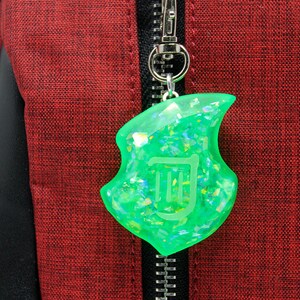 Op de afbeelding: Een groene, glinsterende, harsachtige hanger in de vorm van een noot met een witte omtrek van een harp erin. De hanger is bevestigd aan een zilveren rits.