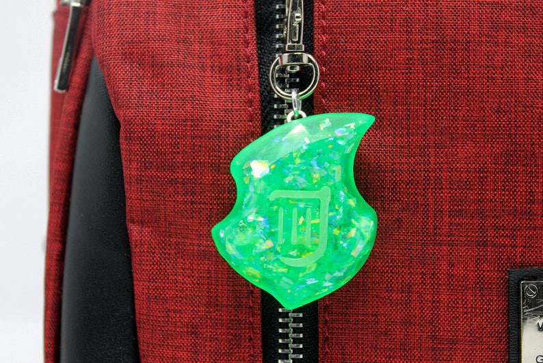 Op de afbeelding: Een groene, glinsterende, harsachtige hanger in de vorm van een noot met een witte omtrek van een harp erin. De hanger is bevestigd aan een zilveren rits.