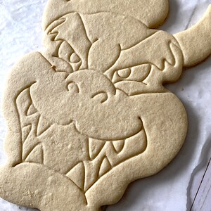 Op de afbeelding: Een eenvoudige, ongedecoreerde suikerkoek in de vorm van een drakenkop. De koek heeft een gedetailleerde omtrek van de drakenkop, inclusief ogen, tanden en een neus.