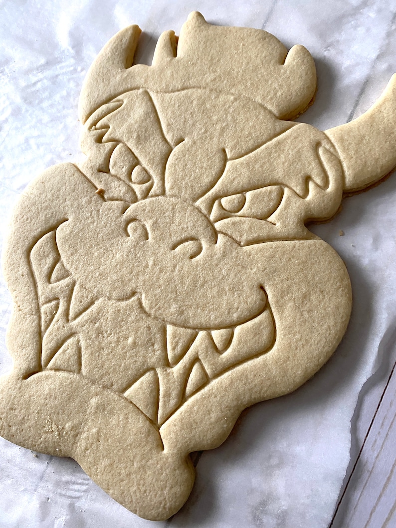 Op de afbeelding: Een eenvoudige, ongedecoreerde suikerkoek in de vorm van een drakenkop. De koek heeft een gedetailleerde omtrek van de drakenkop, inclusief ogen, tanden en een neus.