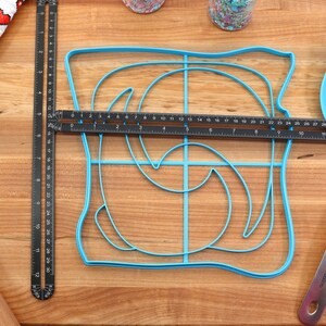 May include: A blue plastic mold shaped like a slice of bread with a swirl design inside. The mold is on a wooden cutting board with a black ruler, a rolling pin, and a blue spatula.