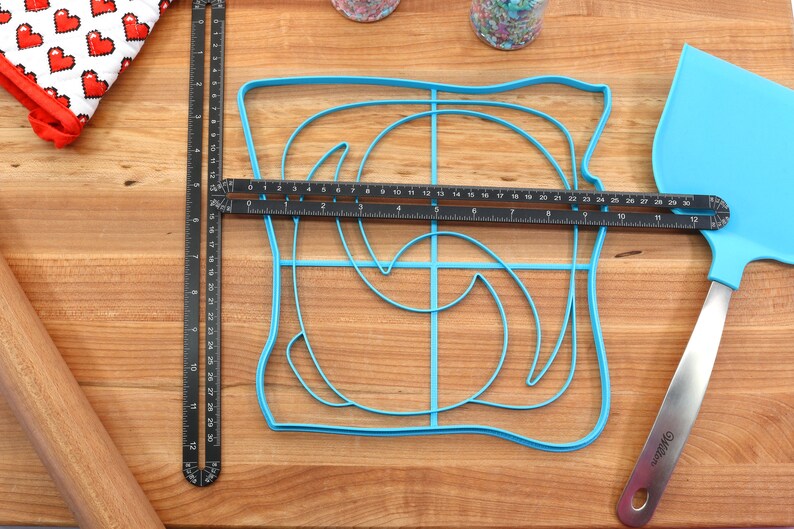 May include: A blue plastic mold shaped like a slice of bread with a swirl design inside. The mold is on a wooden cutting board with a black ruler, a rolling pin, and a blue spatula.