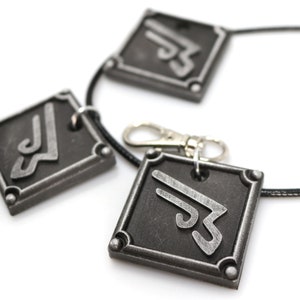 May include: A black necklace with three silver metal pendants. Each pendant is a square with a stylized symbol in the center.