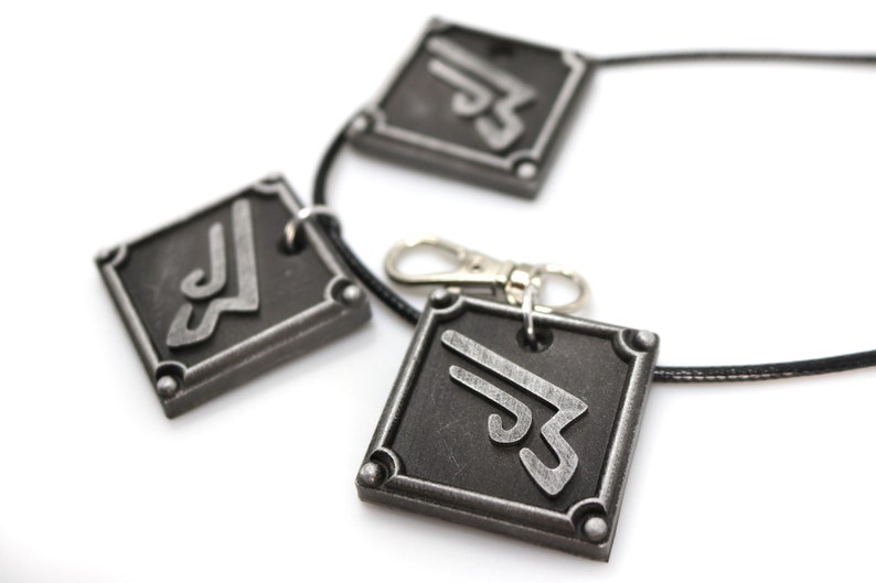 May include: A black necklace with three silver metal pendants. Each pendant is a square with a stylized symbol in the center.