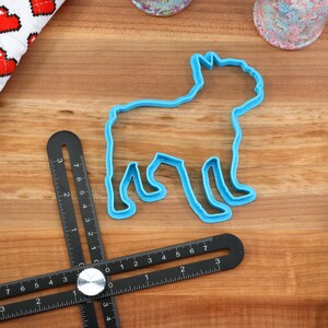 Puede incluir: Un cortador de galletas de pl&aacute;stico azul con forma de perro. El cortador est&aacute; sobre una superficie de madera con una regla negra y una tela blanca y roja con corazones.
