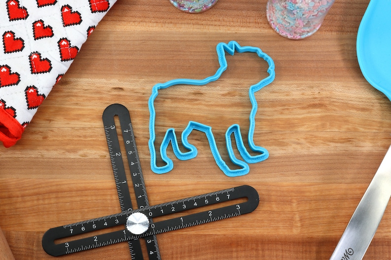 Puede incluir: Un cortador de galletas de pl&aacute;stico azul con forma de perro. El cortador est&aacute; sobre una superficie de madera con una regla negra y una tela blanca y roja con corazones.