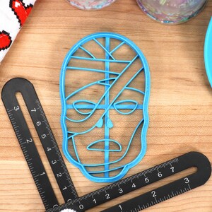 May include: A light blue plastic cookie cutter in the shape of a stylized face. The cutter is surrounded by black measuring tools, a blue spatula, and a white and red heart-patterned cloth. The face design has lines across the eyes and mouth.