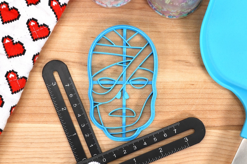 May include: A light blue plastic cookie cutter in the shape of a stylized face. The cutter is surrounded by black measuring tools, a blue spatula, and a white and red heart-patterned cloth. The face design has lines across the eyes and mouth.