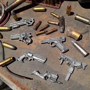 May include: A collection of silver-toned toy guns, including a revolver, a pistol, and a machine gun. The guns are arranged on a brown surface with various bullets and other objects.