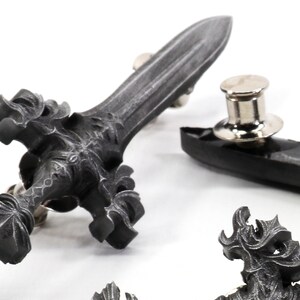May include: A silver-toned sword pin with intricate details. The sword has a pointed tip and a cross-shaped hilt. The pin is attached to a black backing.