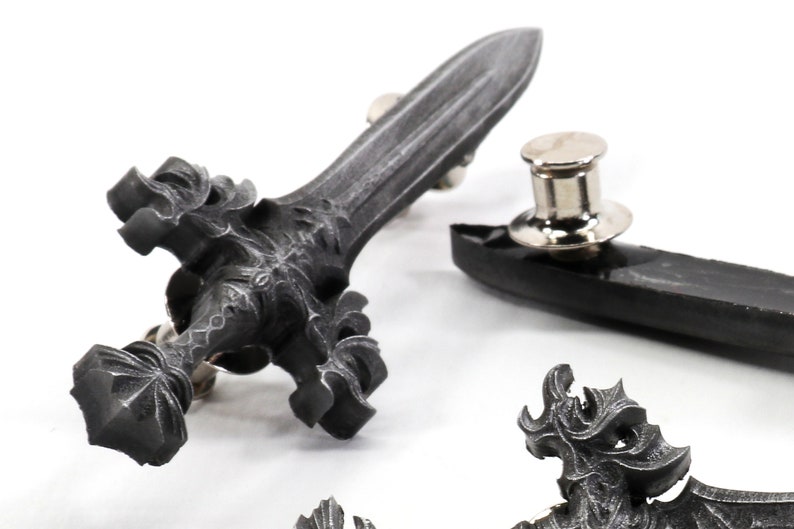 May include: A silver-toned sword pin with intricate details. The sword has a pointed tip and a cross-shaped hilt. The pin is attached to a black backing.