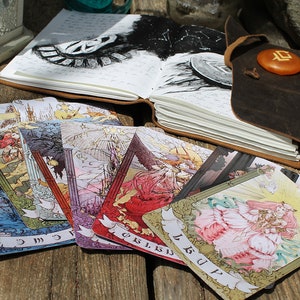 May include: A spread of tarot cards on a wooden table. The cards are illustrated with colorful images and feature a variety of symbols. The cards are arranged in a fan shape, with the top card showing a woman in a pink dress.