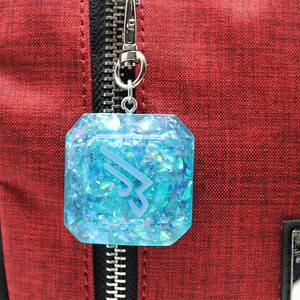 May include: A blue resin charm with a white logo and iridescent glitter, attached to a silver zipper pull. The charm is hanging from a red bag.