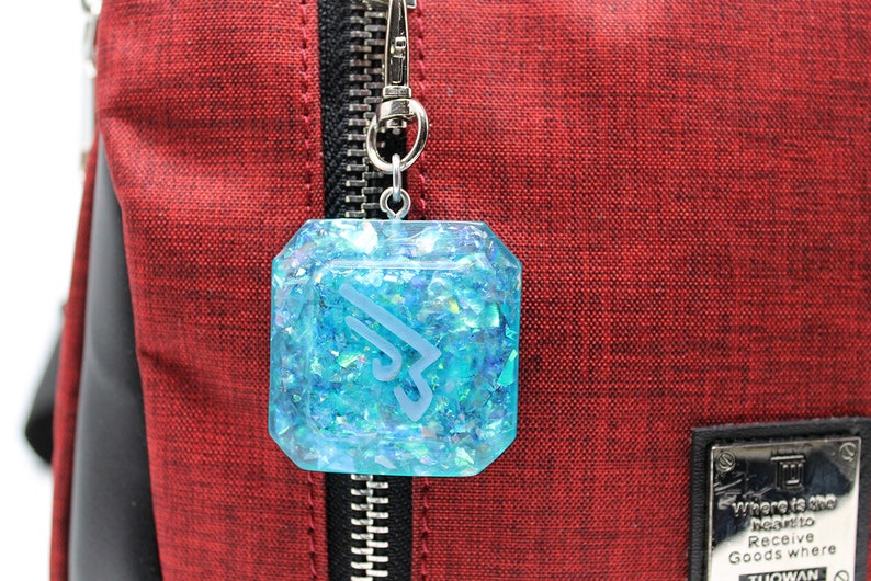 May include: A blue resin charm with a white logo and iridescent glitter, attached to a silver zipper pull. The charm is hanging from a red bag.