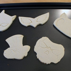 May include: Five unbaked sugar cookies on a black baking sheet. The cookies are shaped like a cat, a mask, a skull, a hat, and a face.