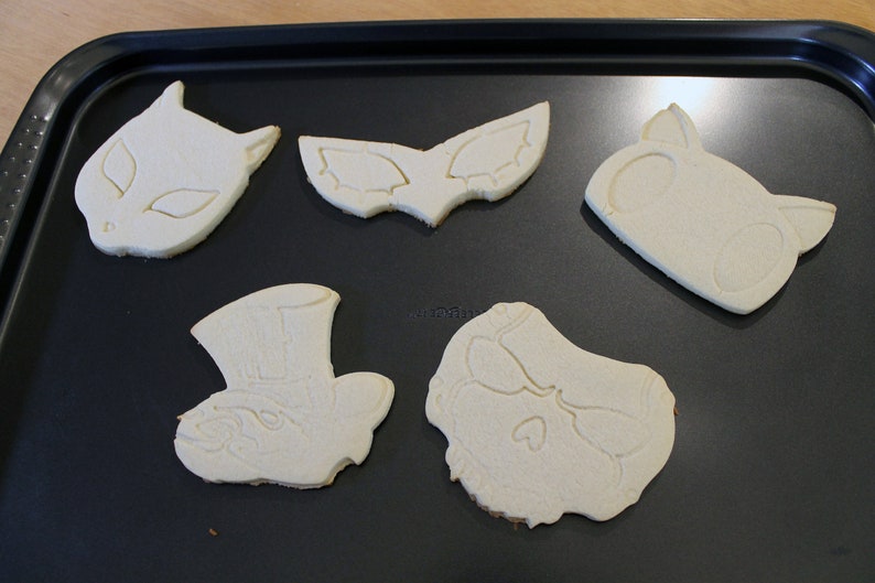 May include: Five unbaked sugar cookies on a black baking sheet. The cookies are shaped like a cat, a mask, a skull, a hat, and a face.