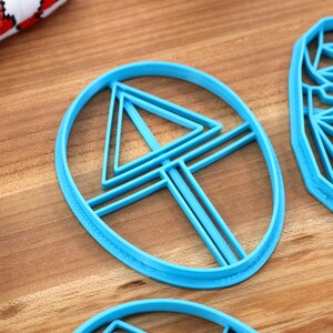 May include: Three blue plastic cookie cutters, one shaped like a circle with a triangle and lines inside, one shaped like a circle with a triangle inside, and one shaped like a face.