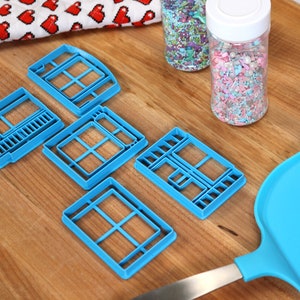 May include: Five blue plastic cookie cutters shaped like windows, a blue spatula, and two jars of colorful sprinkles on a wooden surface.