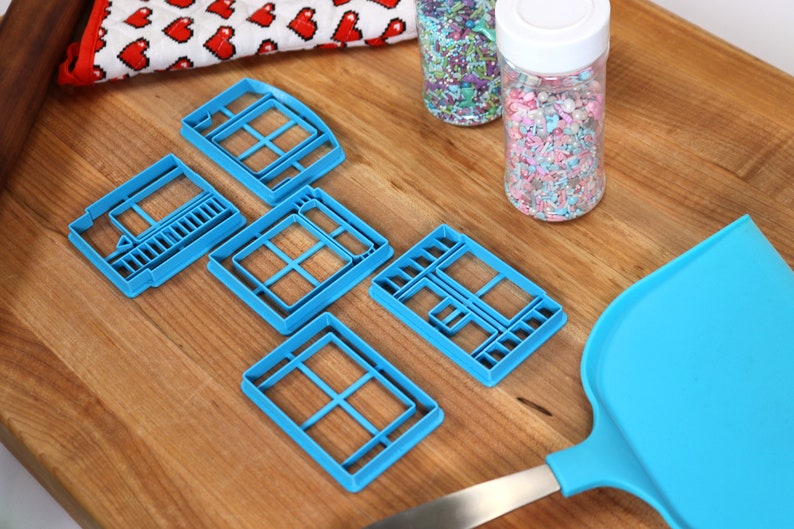 May include: Five blue plastic cookie cutters shaped like windows, a blue spatula, and two jars of colorful sprinkles on a wooden surface.