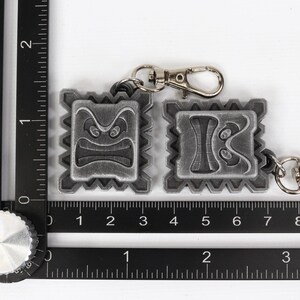 May include: Two silver keychains with a textured, square design. Each keychain features a different cartoon face with a frowning expression. The keychains are attached to silver key rings.