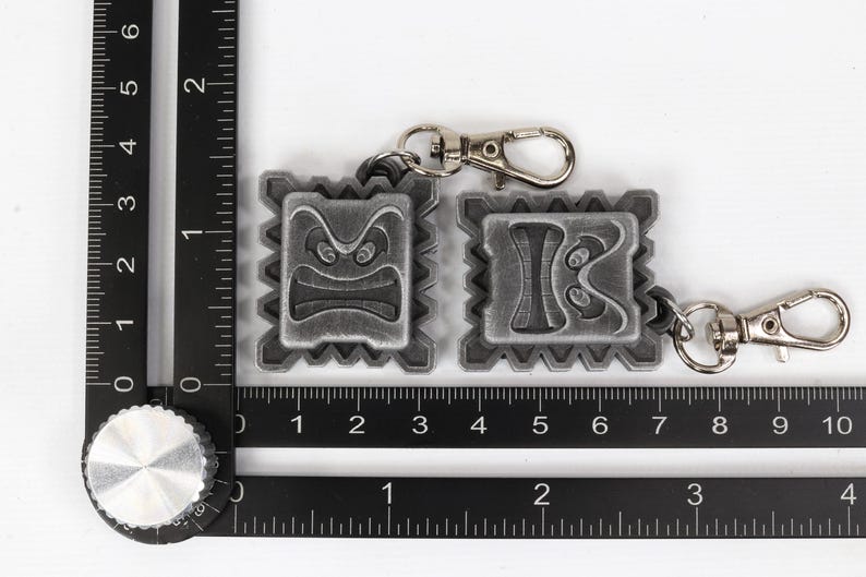 May include: Two silver keychains with a textured, square design. Each keychain features a different cartoon face with a frowning expression. The keychains are attached to silver key rings.