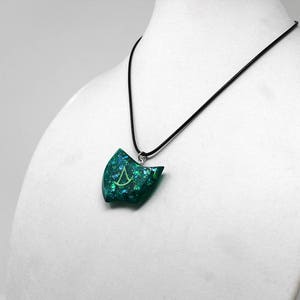 Puede incluir: Un collar con un colgante verde azulado, una cadena plateada y un s&iacute;mbolo verde y plateado dentro del colgante.
