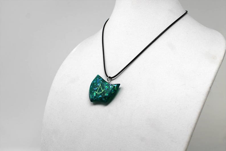Puede incluir: Un collar con un colgante verde azulado, una cadena plateada y un s&iacute;mbolo verde y plateado dentro del colgante.