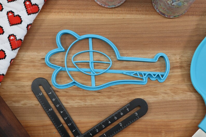 May include: A blue plastic cookie cutter shaped like an alligator with an open mouth and teeth. The cutter has a detailed design with a raised eye and a textured body.
