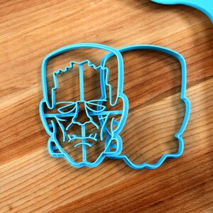 May include: A blue plastic cookie cutter shaped like a Frankenstein's monster face. The cutter is on a wooden surface.