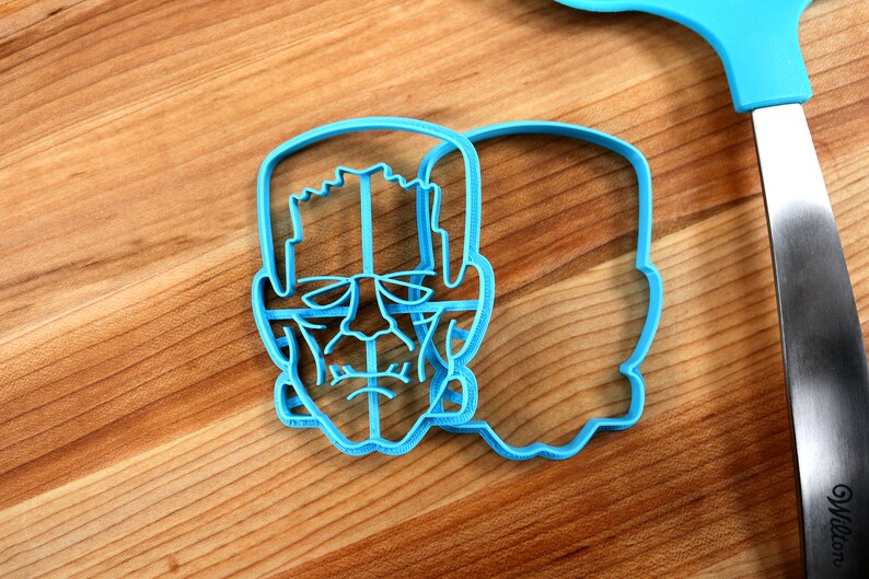 May include: A blue plastic cookie cutter shaped like a Frankenstein's monster face. The cutter is on a wooden surface.