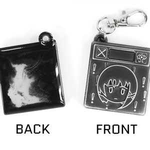 May include: A black and silver metal keychain with a cartoon character on the front and a blank surface on the back. The keychain features a small, silver, metal loop for attaching to a bag or keys.