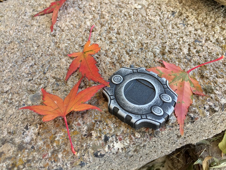 Puede incluir: Un medall&oacute;n de metal plateado con un dise&ntilde;o circular y un centro negro. El medall&oacute;n est&aacute; rodeado de cuatro hojas de arce rojas.
